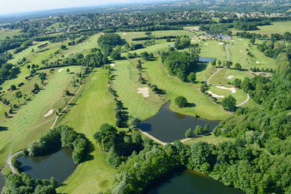Parcours de golf luxuriant vu du ciel. Les fairways sont délimités par des zones boisées, plusieurs étangs et des bunkers clairs.