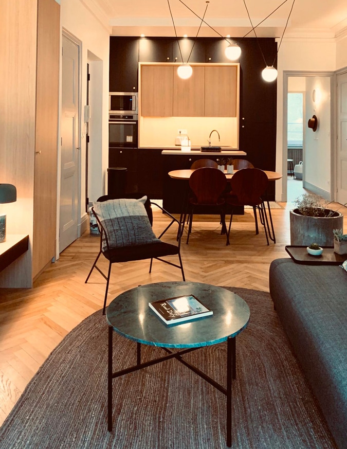 Salon moderne cosy avec parquet en chevrons, table en marbre, fauteuil noir et cuisine ouverte élégante.