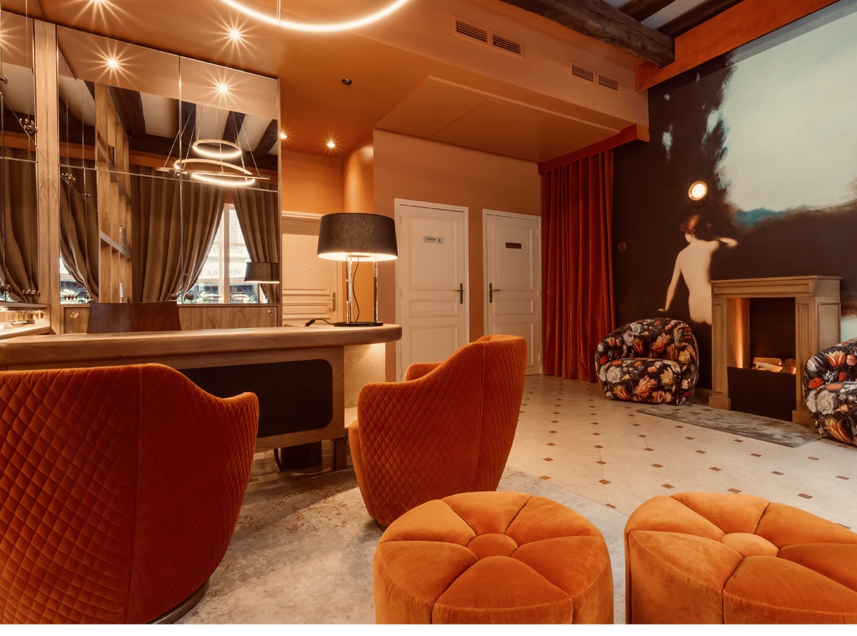 Hall d'hôtel chaleureux : fauteuils matelassés orange et poufs en velours, comptoir en bois, grand miroir. Mural sombre avec cheminée.