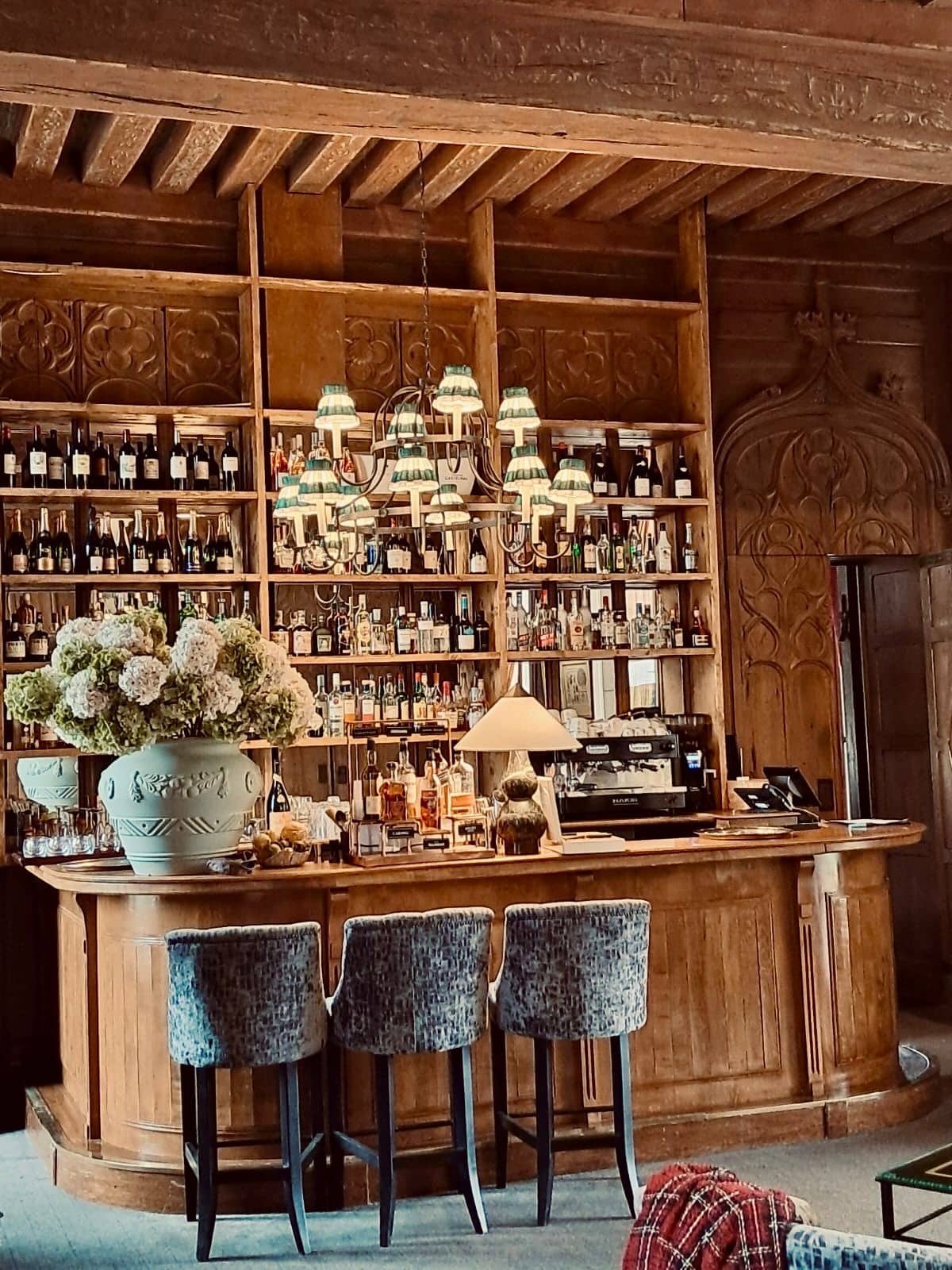 Intérieur historique : bar en bois sculpté et étagères de bouteilles. Vase d'hydrangeas, tabourets luxueux et plafond à poutres.