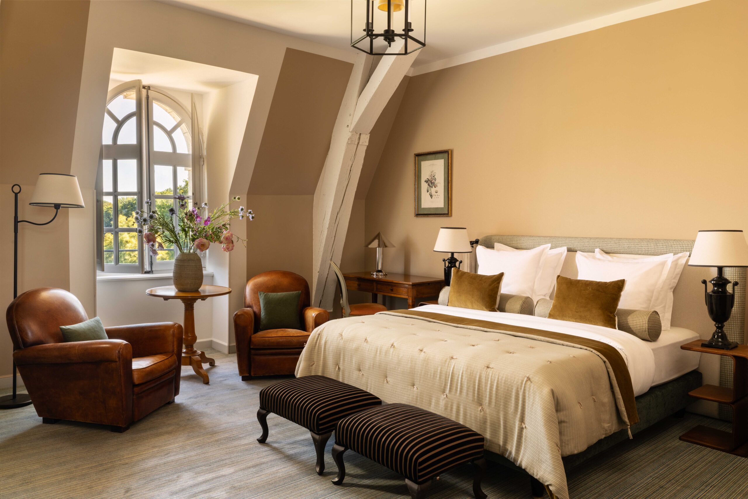 Suite mansardée luxueuse, lit double, fauteuils club en cuir, table de chevet antique, poutres blanchies et fenêtre arquée.