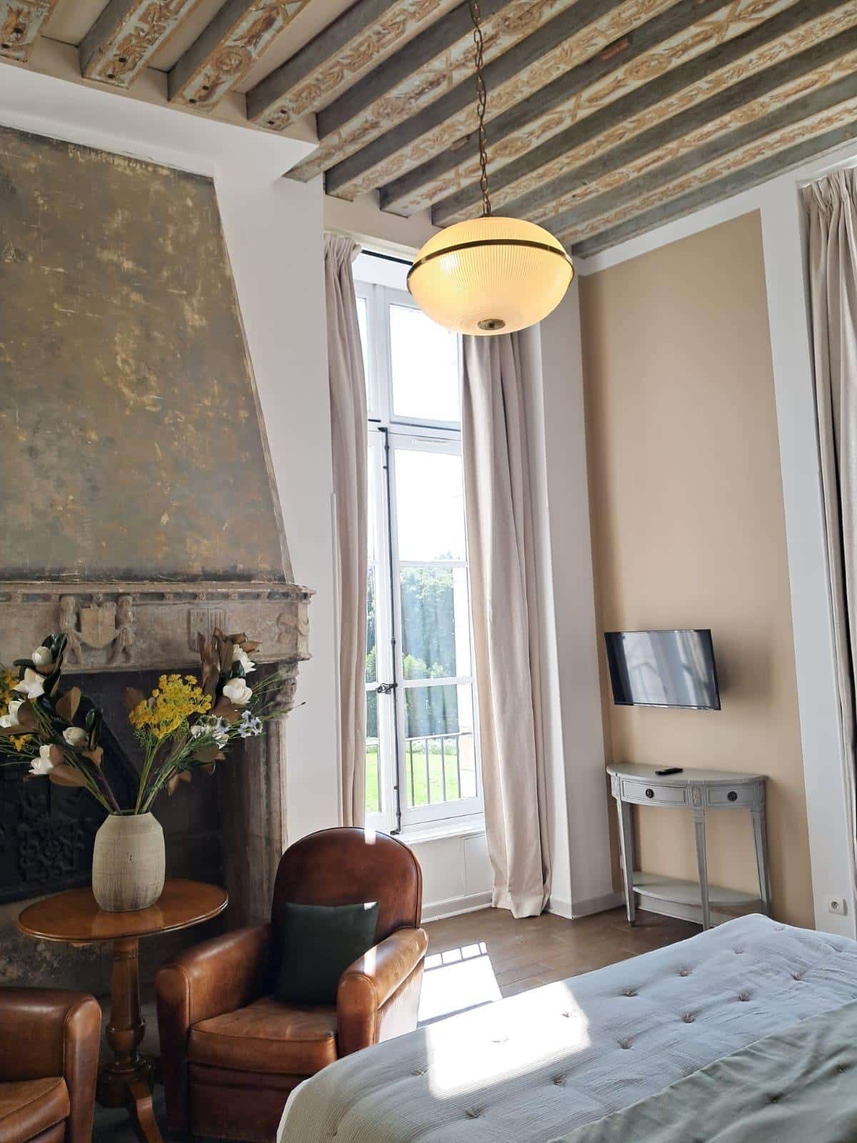 Luxueuse chambre rustique chic: poutres apparentes, cheminée en pierre ancienne, fauteuil club en cuir et lumière naturelle près de la fenêtre jardin.