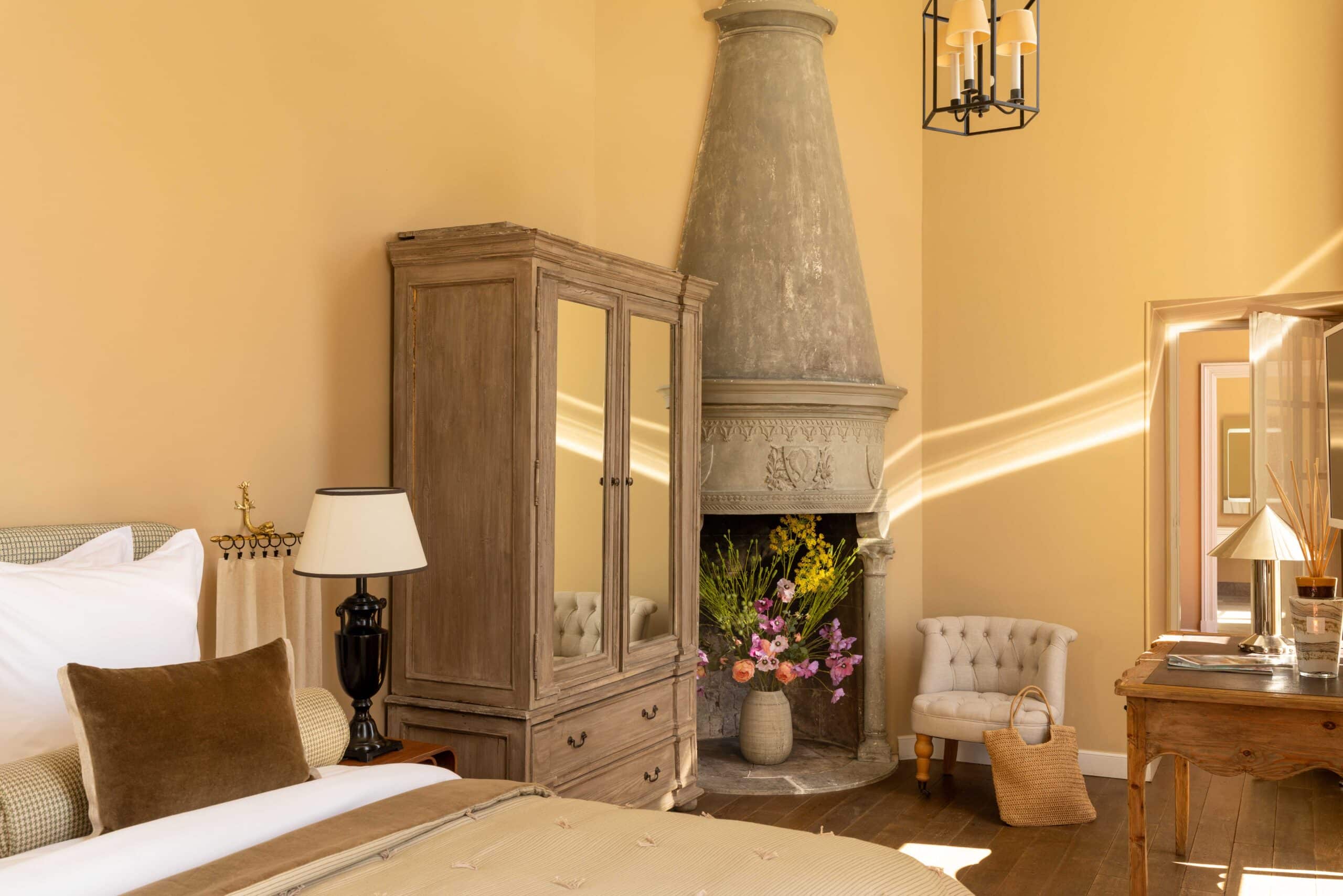 Intérieur chaleureux: chambre d'hôtel élégante avec murs ocre, armoire miroir, et grande cheminée fleurie. Rayons de soleil.