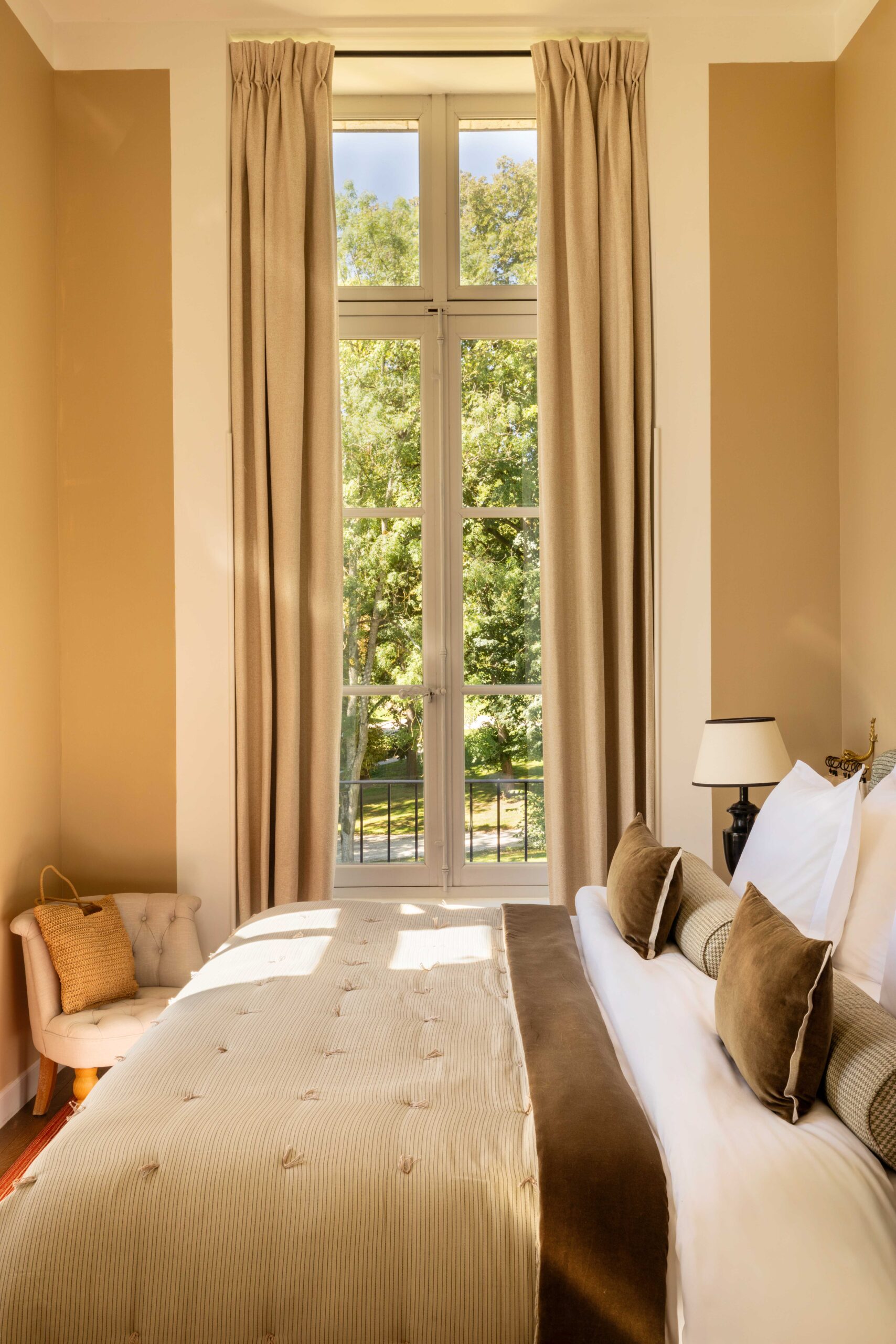 Intérieur de suite hôtel luxueuse. Grand lit, coussins marron et rideaux beige devant une fenêtre haute s'ouvrant sur un jardin verdoyant.