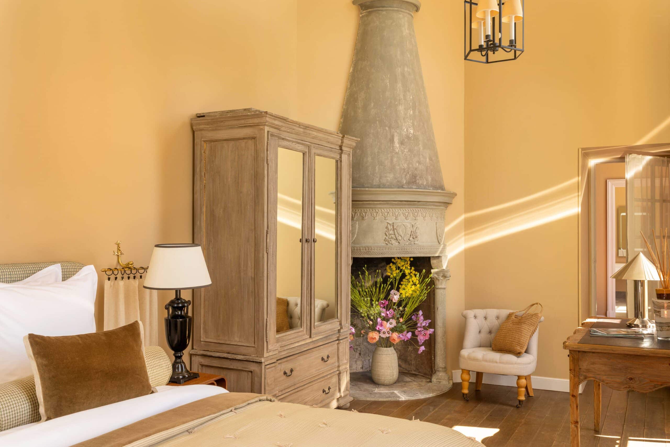 Intérieur élégant : murs ocre, armoire ancienne à miroir, et cheminée rustique en pierre. Lumière chaude du soleil sur un lit douillet et des fleurs.