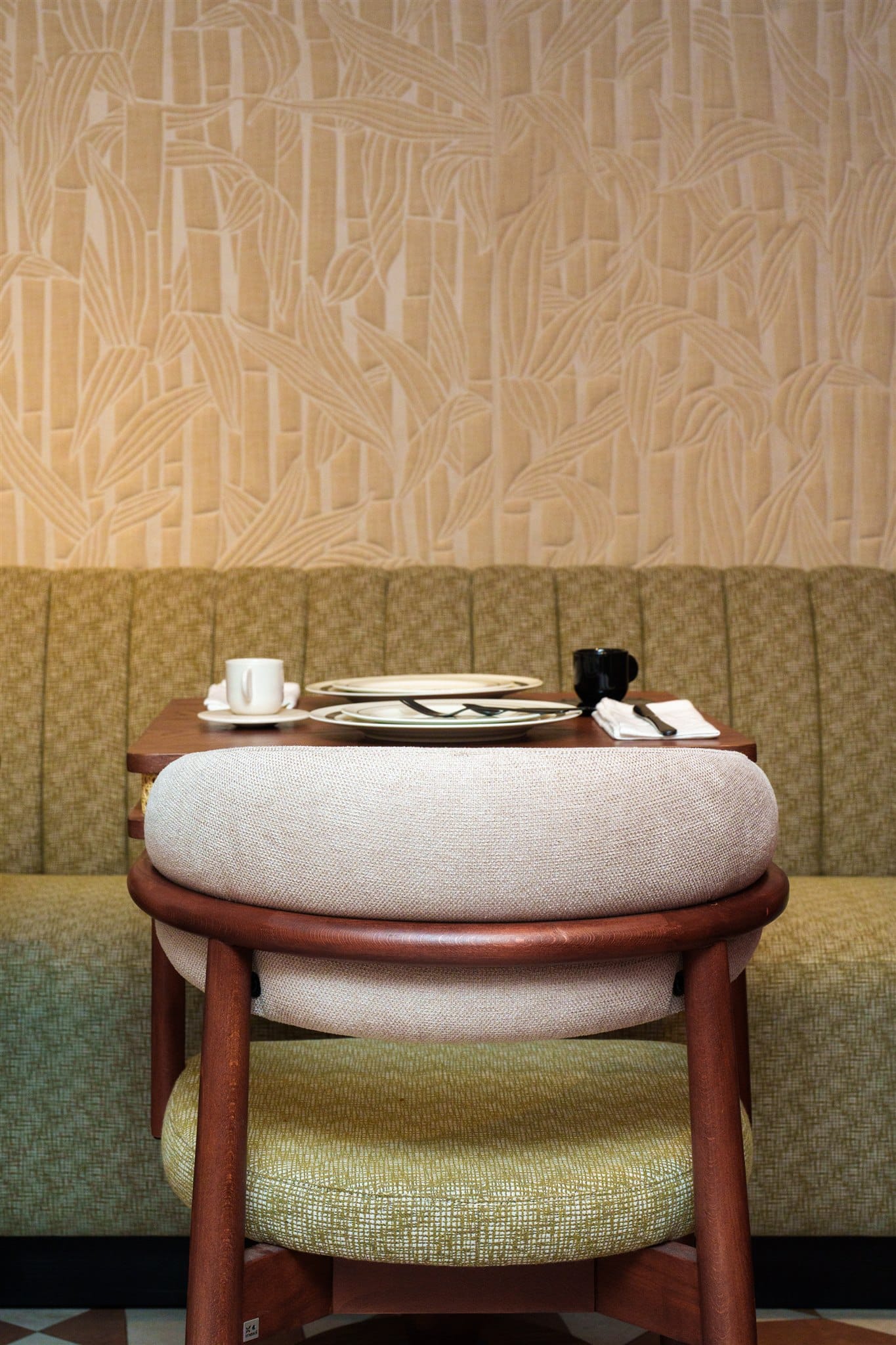 Chaise en bois et banquette verte dans un intérieur de restaurant. Le mur présente un papier peint texturé beige à motif de bambou. Table dressée.