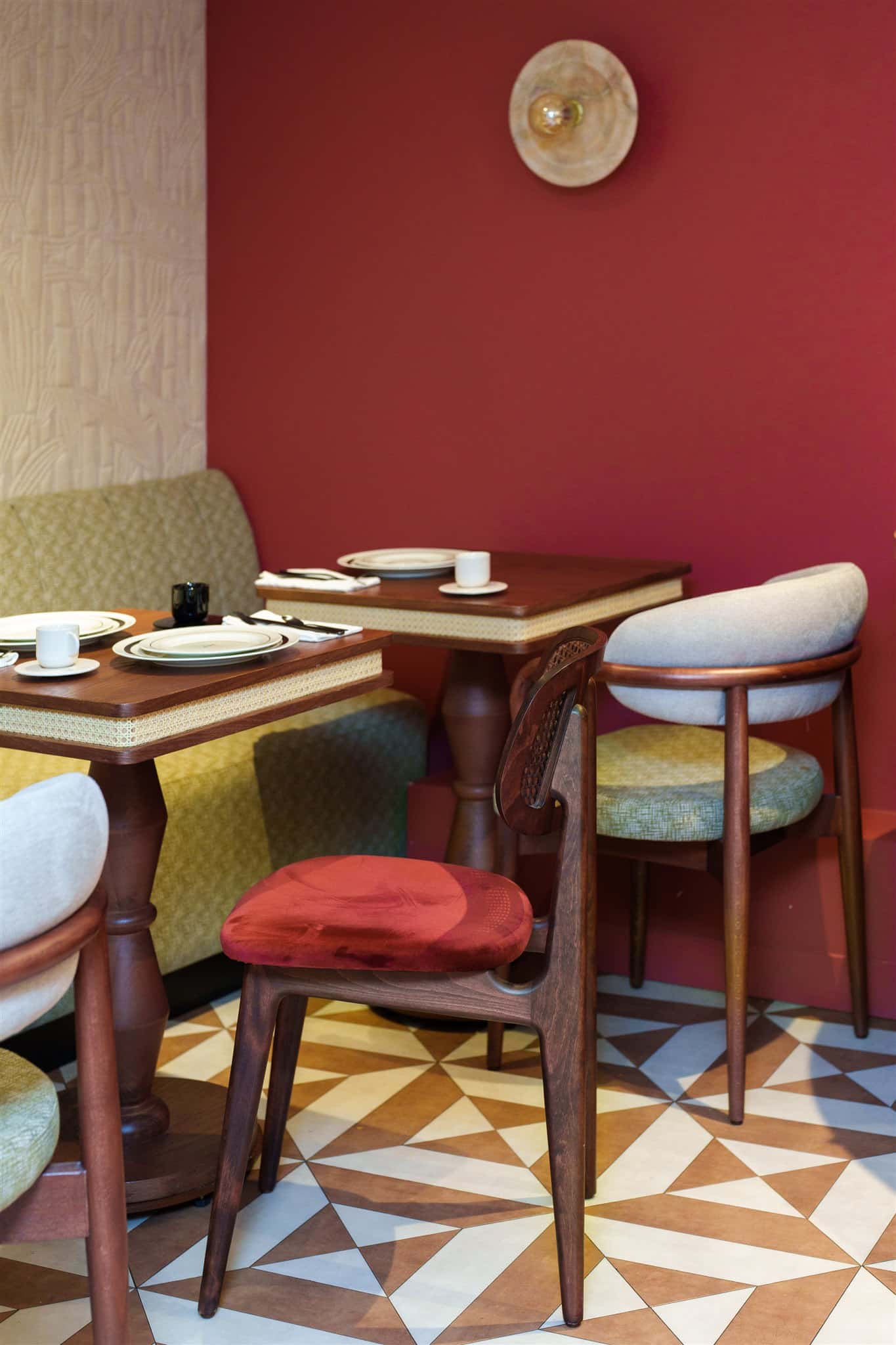 Salle de restaurant chaleureuse: murs rouge bordeaux, tables en bois canné, banquette vert olive et chaises design sur carrelage géométrique.