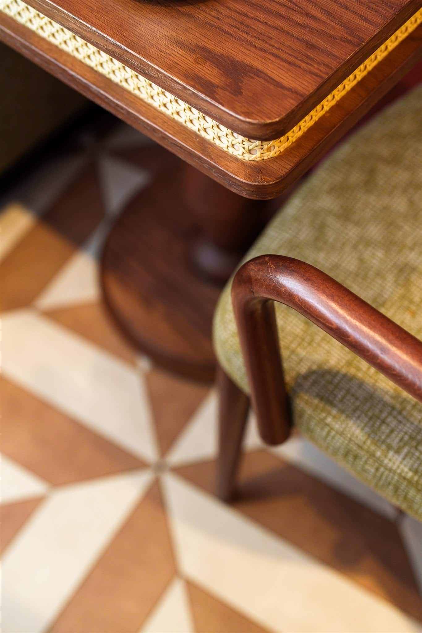 Coin de table en bois sombre avec insert en cannage et accoudoir courbé de chaise verte. Sol carrelé géométrique beige et terre cuite.
