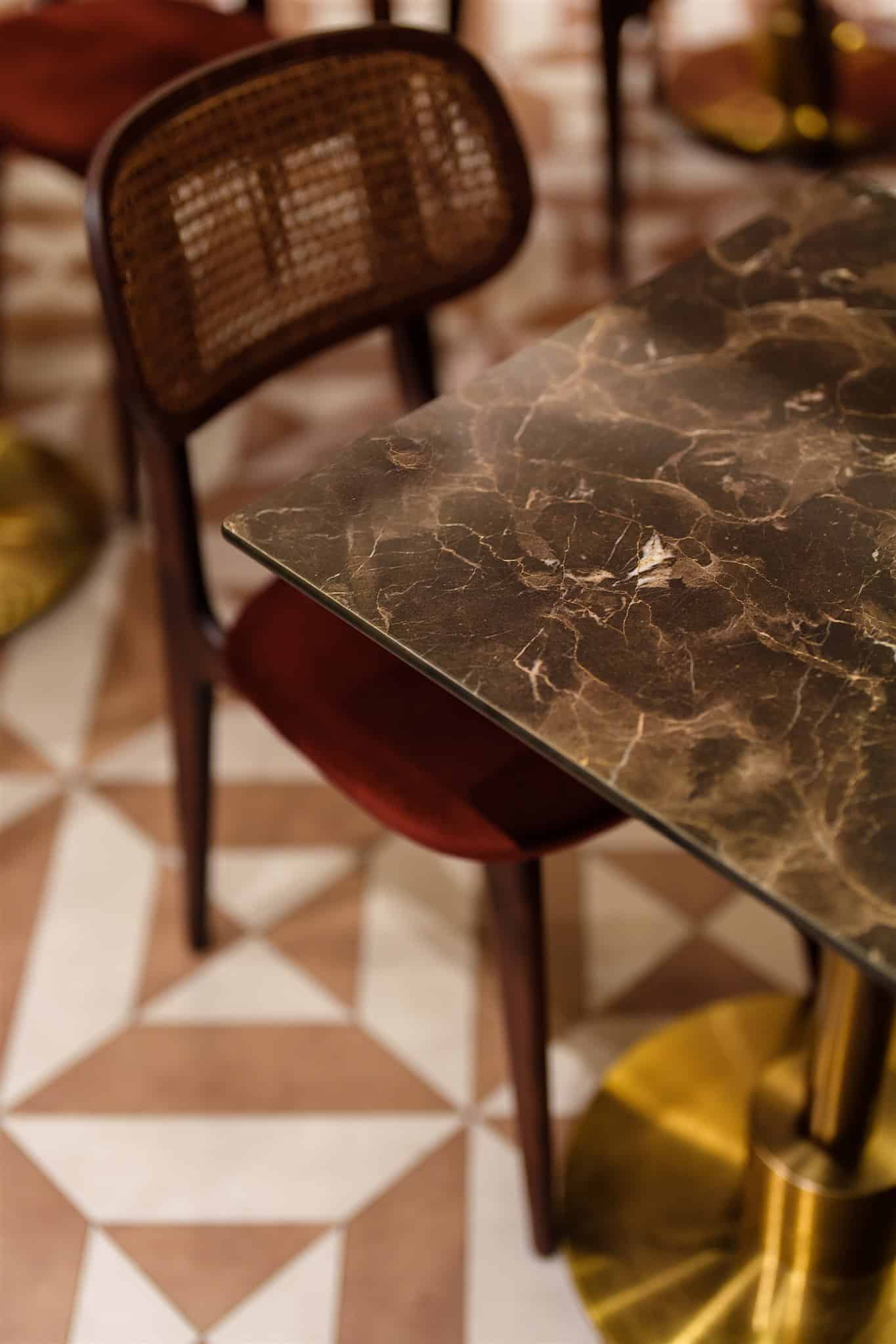 Table marbre foncé veiné, pied en laiton doré, et chaise rotin à assise velours rouge. Sol à motifs géométriques marron clair.