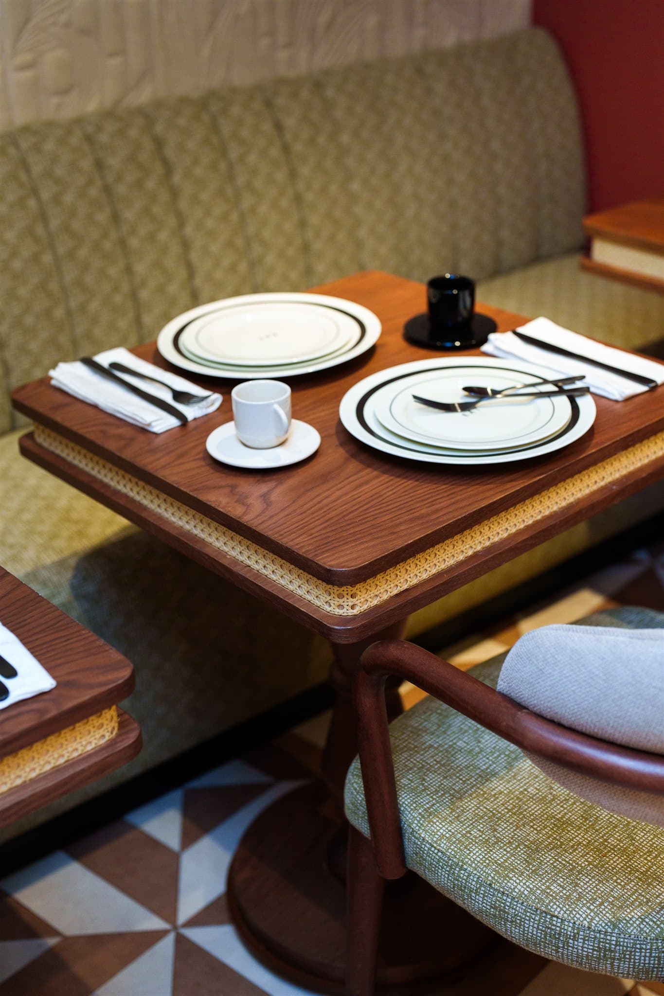 Élégante table en bois et cannage prête pour le service. Deux mises en place avec vaisselle blanche et banquette texturée verte.